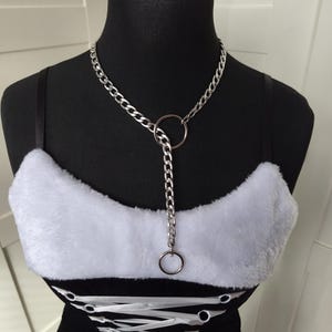 Könnte beinhalten: Ein schwarzes Bustier mit weißem Kunstfellbesatz und einer silbernen Halskette. Die Halskette hat einen großen Ring und eine herabhängende Kette mit einem kleineren Ring. Das Bustier hat eine Schnürung vorne.