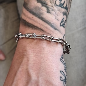 Könnte beinhalten: Ein silberfarbenes Armband mit Kettenglied-Design. Das Armband hat miteinander verbundene Glieder mit kleinen, runden Kugeln an den Verbindungsstellen. Der Schmuck wird an einem Handgelenk mit sichtbaren Tätowierungen getragen.