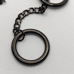 Könnte beinhalten: Nahaufnahme eines schwarzen Metall-Schlüsselanhängers mit zwei großen Ringen und einer Kette. Die Ringe sind durch eine Kette aus kleineren Gliedern verbunden. Das Metall hat eine glänzende, reflektierende Oberfläche.