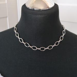 Könnte beinhalten: Eine silberfarbene Kettenhalskette bestehend aus einer Reihe sich wiederholender ovaler Glieder, die durch kleine, runde Metallperlen verbunden sind. Die Kette wird auf einer schwarzen Schaufensterpuppe präsentiert und betont das metallische Finish.