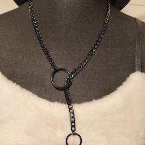 Könnte beinhalten: Eine schwarze Metallkettenhalskette mit Y-förmigem Design und zwei kreisförmigen Ringen. Die Kettenglieder sind mit einem größeren Ring in der Mitte verbunden, der nach unten zu einem kleineren Ring am Ende der Halskettenstruktur verläuft.
