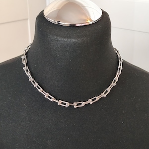 Könnte beinhalten: Eine silberfarbene Halskette mit U-förmigem Kettenglied-Design wird auf einer schwarzen Schaufensterpuppe präsentiert. Die Halskette ist im Choker-Stil gehalten, und die Glieder sind gleichmäßig verteilt. Die Schaufensterpuppe hat eine silberne Kuppel oben.