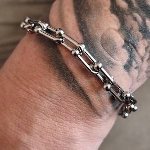 Könnte beinhalten: Ein silberfarbenes Metallkettenarmband mit einer Reihe länglicher, rechteckiger Glieder, die an jeder Verbindungsstelle durch kleine, polierte, kugelförmige Perlen verbunden sind und ein strukturiertes, geometrisches Muster bilden.