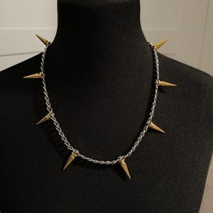 Könnte beinhalten: Eine silberfarbene Halskette mit goldfarbenen Stacheln. Die Halskette wird auf einer schwarzen Schaufensterpuppe präsentiert. Die Stacheln sind gleichmäßig um die Kette verteilt. Ein auffälliges Accessoire.