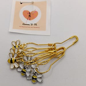 Könnte beinhalten: Goldfarbene Sicherheitsnadeln mit dekorativen Bienen-Anhängern. Die Bienen haben weiße, gelbe und schwarze Emaille-Details. Ein kleines Etikett mit dem Text "Aurora's S-M Handgemachtes aus Hamburg" ist ebenfalls vorhanden.