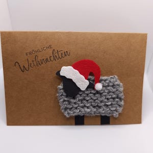 Könnte beinhalten: Eine braune Karte mit dem Text "Fröhliche Weihnachten" und einem grauen Schaf mit rotem und weißem Weihnachtsmütze. Das Schaf hat schwarze Beine und ein schwarzes Gesicht.
