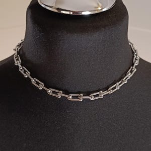 Könnte beinhalten: Eine silberfarbene Halskette mit rechteckigen Gliedern und kleinen, runden Kugeln. Die Halskette wird auf einer schwarzen Schaufensterpuppe präsentiert, um ihr Design zu zeigen. Der Schmuck ist im Choker-Stil gehalten.