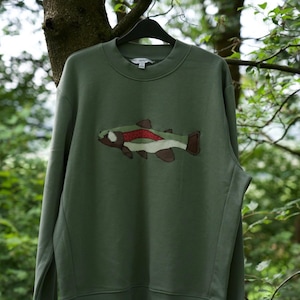 May include: An olive green sweatshirt with an embroidered fish design in brown, red, and white. The crew neck top is suspended on a black hanger, set against a backdrop of green trees. A casual, nature-themed garment.