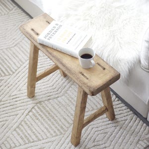 Peut inclure: Un tabouret en bois avec une finition naturelle et un livre intitulé "12 Rules for Life" de Jordan B. Peterson posé dessus. Une tasse à café blanche est posée sur le livre.