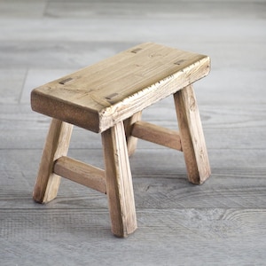 May include: A rustic wooden stool with a rectangular top and three legs. The stool is made of natural wood and has a weathered finish.