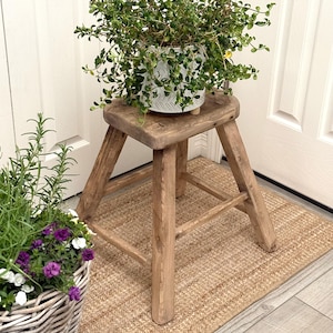 May include: A small, weathered wooden stool with a square top and three legs. A white, patterned ceramic pot with greenery is on top. A woven basket with purple and white flowers is in the foreground, and a jute rug is beneath the stool.