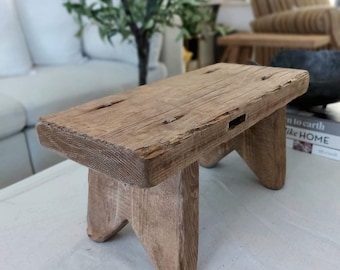 May include: A rustic wooden stool with a rectangular top and four legs. The stool is made of natural wood and has a weathered finish.