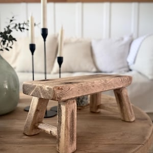 May include: A small, rectangular wooden stool with a natural, weathered finish. The stool features four sturdy, thick legs connected by horizontal support bars on the sides, showcasing a visible wood grain texture and rustic, unfinished edges.