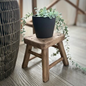 May include: A small, weathered wooden stool supports a black plant pot filled with trailing greenery. The stool has a square top and four angled legs. A woven basket is partially visible on the left. The scene is lit with natural light, creating a warm atmosphere.