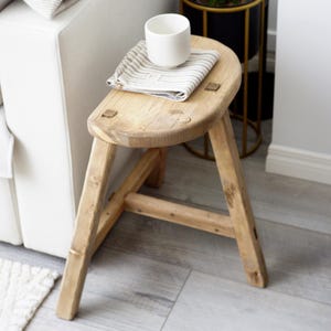 May include: A small, rustic wooden stool with a round seat and three angled legs. The stool is made of light-colored wood and has visible wood grain. A white cup and a striped cloth are on the seat.