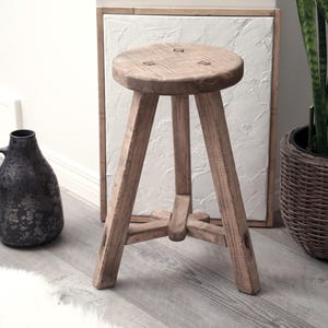 May include: A rustic wooden stool with a triangular base and a natural wood finish. The stool is made of solid wood and has a simple, farmhouse style.