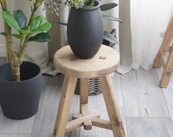 May include: A rustic wooden stool with three legs and a round top. A black ceramic vase with a plant is sitting on the stool.