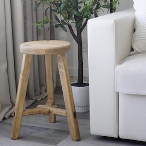 Peut inclure: Un tabouret en bois avec un siège rond et trois pieds. Le tabouret est en bois naturel et a une finition rustique. Il est placé dans un salon à côté d'un canapé blanc et d'une plante en pot.