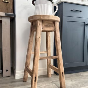 May include: A tall, round wooden stool with a light brown, weathered finish. The stool features four straight legs connected by horizontal support bars. A white, ribbed ceramic pitcher containing a green plant sits on the flat, circular seat.