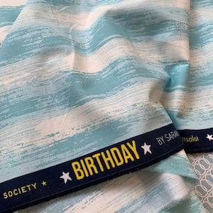 Könnte beinhalten: Blau-weißer Stoff mit einem Pinselstrich-Design. Der Stoff hat einen marineblauen Rand mit dem Wort "BIRTHDAY" in gelben Buchstaben und weißen Sternen. Der Stoff ist von Ruby Star Society.