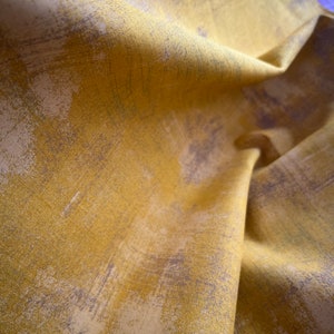May include: Close-up of folded fabric in mustard yellow with abstract brushstroke patterns in grey and white. The fabric has a textured appearance and a slight sheen, suggesting a high-quality material. The folds create shadows.
