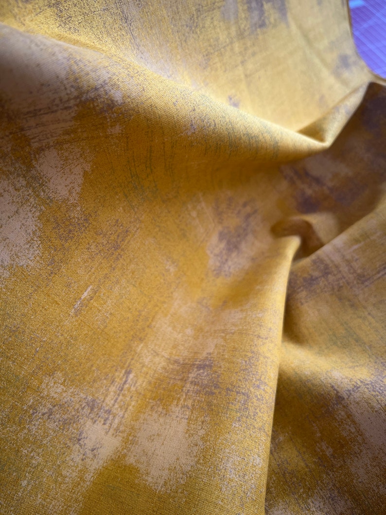 May include: Close-up of folded fabric in mustard yellow with abstract brushstroke patterns in grey and white. The fabric has a textured appearance and a slight sheen, suggesting a high-quality material. The folds create shadows.