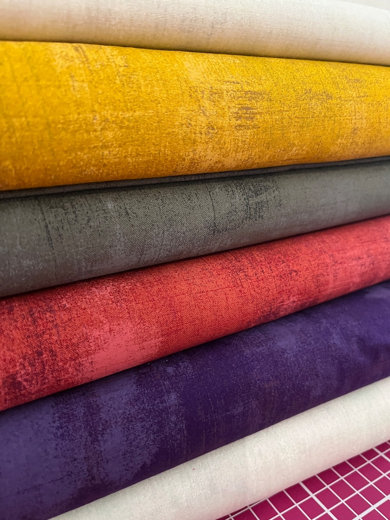 May include: A stack of six fabric rolls in various colors, including beige, yellow, gray, red, purple, and beige. The fabric has a textured, distressed look.