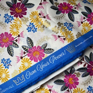 Könnte beinhalten: Ein weißer Stoff mit einem floralen Muster in den Farben Rosa, Gelb, Blau und Gold. Der Stoff hat ein blaues Band mit dem Text "Own Your Power!" und eine goldene Krone.