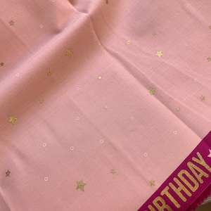 Könnte beinhalten: Rosafarbener Stoff mit einem goldenen Sternenmuster und einem rosa Band mit dem Wort "Birthday" in goldenen Buchstaben.