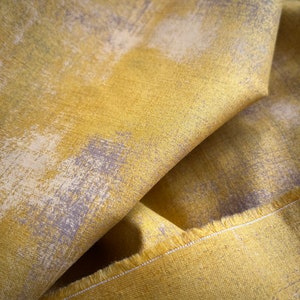 May include: Close-up of folded fabric with a textured surface. The fabric is mainly yellow with a pattern of light grey and off-white brushstrokes. The edge of the fabric is visible, showing a raw, unfinished edge.