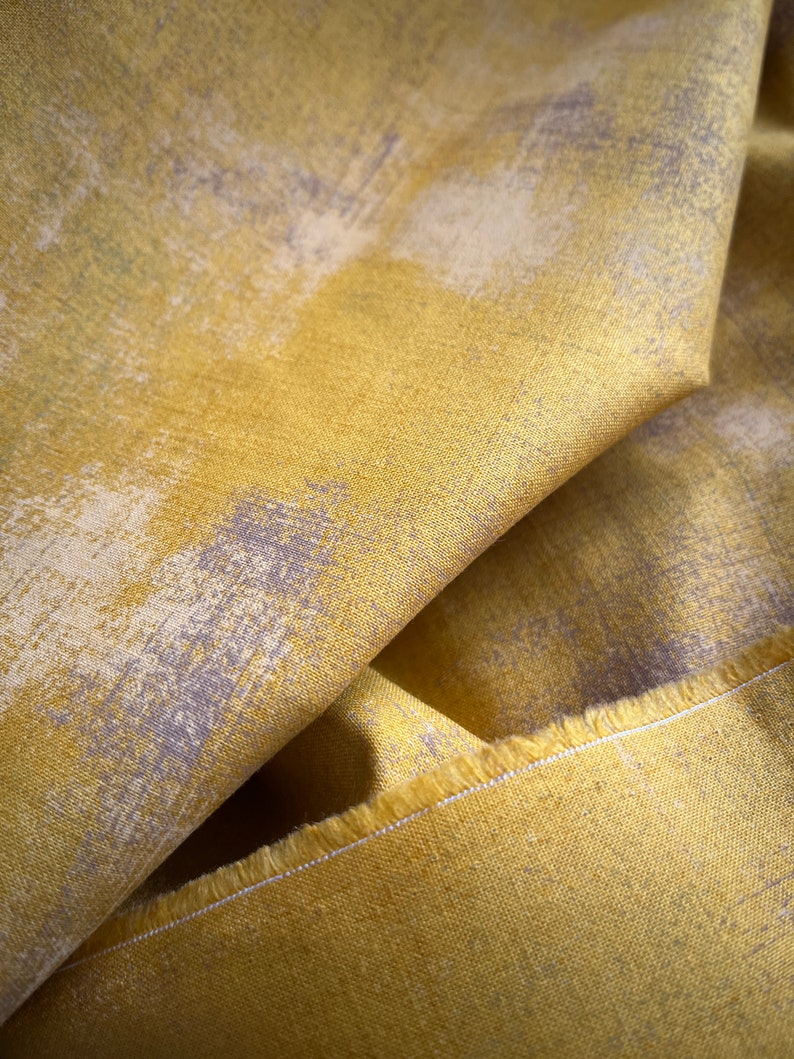 May include: Close-up of folded fabric with a textured surface. The fabric is mainly yellow with a pattern of light grey and off-white brushstrokes. The edge of the fabric is visible, showing a raw, unfinished edge.
