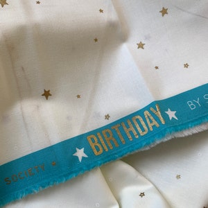 Könnte beinhalten: Weißer Stoff mit einem goldenen Sternmuster. Ein türkisfarbenes Band mit dem Text "SOCIETY BIRTHDAY BY" ist am unteren Rand des Stoffes angenäht.
