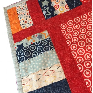 Könnte beinhalten: Ein Patchwork-Quilt mit rotem Rand und verschiedenen Stoffstreifen mit Mustern. Die Stoffe zeigen Sterne, Kreise und gepunktete Designs in Blau-, Orange- und Weißtönen. Der Quilt ist mit einem hellgrauen Stoff eingefasst.