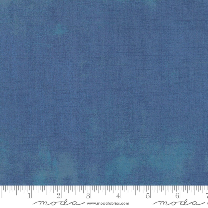 May include: A close-up of a blue fabric with a textured appearance. The fabric has a mottled effect with lighter blue and teal accents. A ruler is at the bottom of the image, with the word "moda" printed on it.