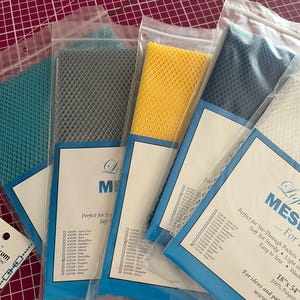 Könnte beinhalten: Mehrere Mesh-Stoffmuster in verschiedenen Farben, darunter Türkis, Grau, Gelb und Marineblau, werden in transparenten Plastiktüten präsentiert. Jede Tüte hat ein blaues Etikett mit Text und einer Farbliste. Das Mesh ist 45.7cm x 137.2cm groß.