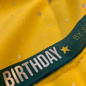 Könnte beinhalten: Gelber Stoff mit einem grünen Band, auf dem "BIRTHDAY" in silbernen Buchstaben steht. Der Stoff ist mit kleinen weißen Kreisen und silbernen Sternen bedeckt.