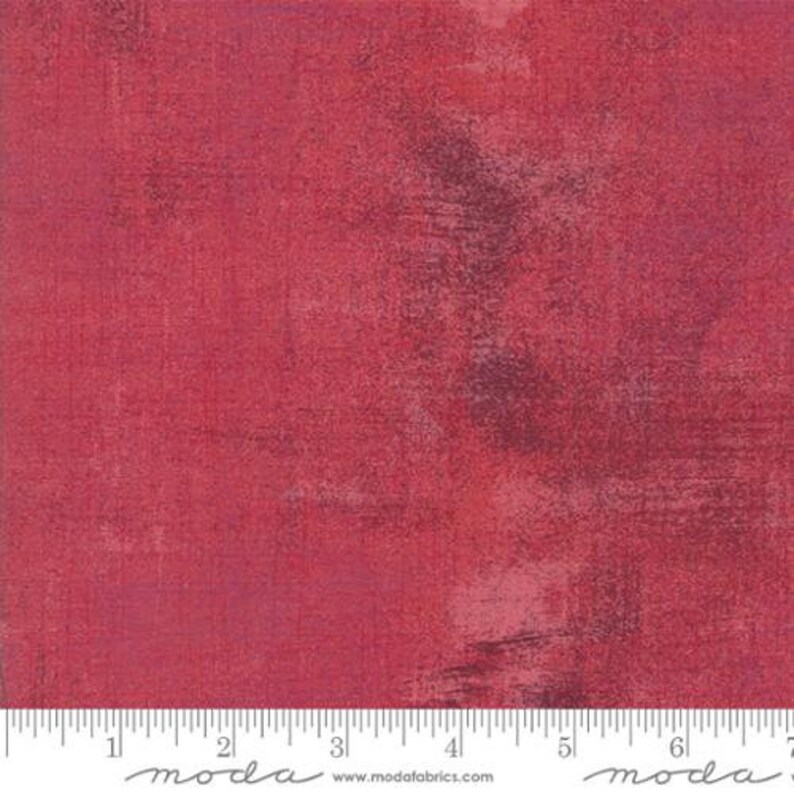 May include: Red fabric with a textured, distressed look. The fabric has a subtle, mottled pattern with shades of pink and purple. The fabric is shown with a ruler for scale.