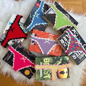 May include: A collection of seven handmade crochet book sleeves shaped like thong underwear, each featuring a small ribbon bow at the centre. The colours include red, light blue, lime green, lavender, hot pink, yellow, and a multi-coloured pattern.