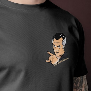 Può includere: T-shirt grigio scuro con un'illustrazione a cartone animato di un uomo con i capelli all'indietro che indica con il dito. L'illustrazione è sull'area del petto sinistro. La maglietta è in materiale morbido.