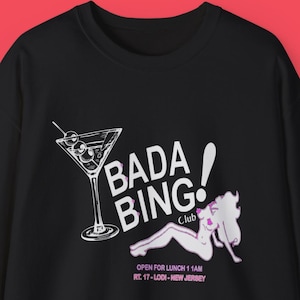 Può includere: Felpa nera con una grafica raffigurante un bicchiere da martini e la scritta "Bada Bing! Club". Una figura dei cartoni animati è sdraiata sotto il testo. La felpa include anche la scritta "Open for Lunch 11 AM RT. 17-Lodi-New Jersey".