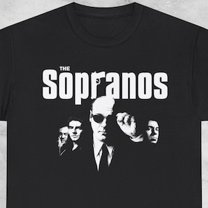 Può includere: T-shirt nera con la scritta bianca "The Sopranos" sopra un'immagine in bianco e nero di diverse persone. L'immagine è una grafica promozionale per la serie televisiva.