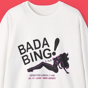 Può includere: Felpa bianca con la scritta "BADA BING!" in nero e una grafica di una donna in nero e rosa. La felpa include anche il testo "OPEN FOR LUNCH 11AM RT. 17 - LODI - NEW JERSEY".