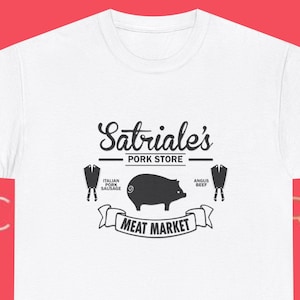 Könnte beinhalten: Weißes T-Shirt mit einer schwarzen Grafik eines Schweins und dem Text "Satriale's Pork Store Meat Market". Das Design beinhaltet Illustrationen von Würstchen und Rindfleischstücken. Das Shirt besteht aus einem weichen, bequemen Material.
