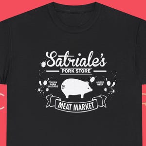 Könnte beinhalten: Schwarzes T-Shirt mit einem weißen Schweine-Grafik-Design und dem Text "Satriale's Pork Store Meat Market". Das Design enthält auch die Texte "Italian Pork Sausage" und "Angus Beef".