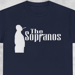 Può includere: T-shirt blu navy con una silhouette bianca di un uomo che fuma una sigaretta e la scritta "The Sopranos" in bianco. Il design è centrato sulla parte anteriore della maglietta, creando un effetto sorprendente.