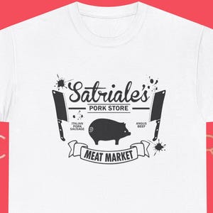 Könnte beinhalten: Weißes T-Shirt mit schwarzem Grafikdesign. Das Design zeigt den Text "Satriale's Pork Store" über einer Schweineillustration, darunter "Meat Market". Zwei Hackmesser flankieren das Schwein.