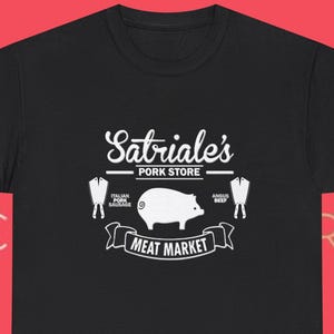 Könnte beinhalten: Schwarzes T-Shirt mit weißer Grafik, das ein Schwein, die Worte "Meat Market" und "Satriale's Pork Store" zeigt. Das Design enthält Illustrationen von Würsten und Fleischstücken. Das T-Shirt hat einen Rundhalsausschnitt.