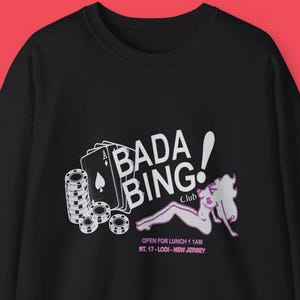 Può includere: Felpa nera con la scritta "Bada Bing! Club" e una grafica di carte da gioco, fiches da poker e una figura reclinata. La felpa include anche la scritta "Open for Lunch 11 AM RT. 17 - Lodi - New Jersey."