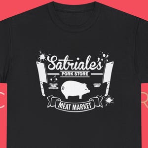 Könnte beinhalten: Schwarzes T-Shirt mit weißer Grafik. Das Design zeigt den Text "Satriale's Pork Store", eine Schweine-Illustration und die Worte "Meat Market" auf einem Banner. Zwei Hackmesser sind ebenfalls abgebildet.