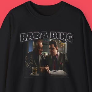 Può includere: Felpa girocollo nera con una grafica di due uomini seduti a un tavolo, con la scritta "Bada Bing Club" sopra. L'immagine è ambientata su uno sfondo rosso. La felpa ha le maniche lunghe.